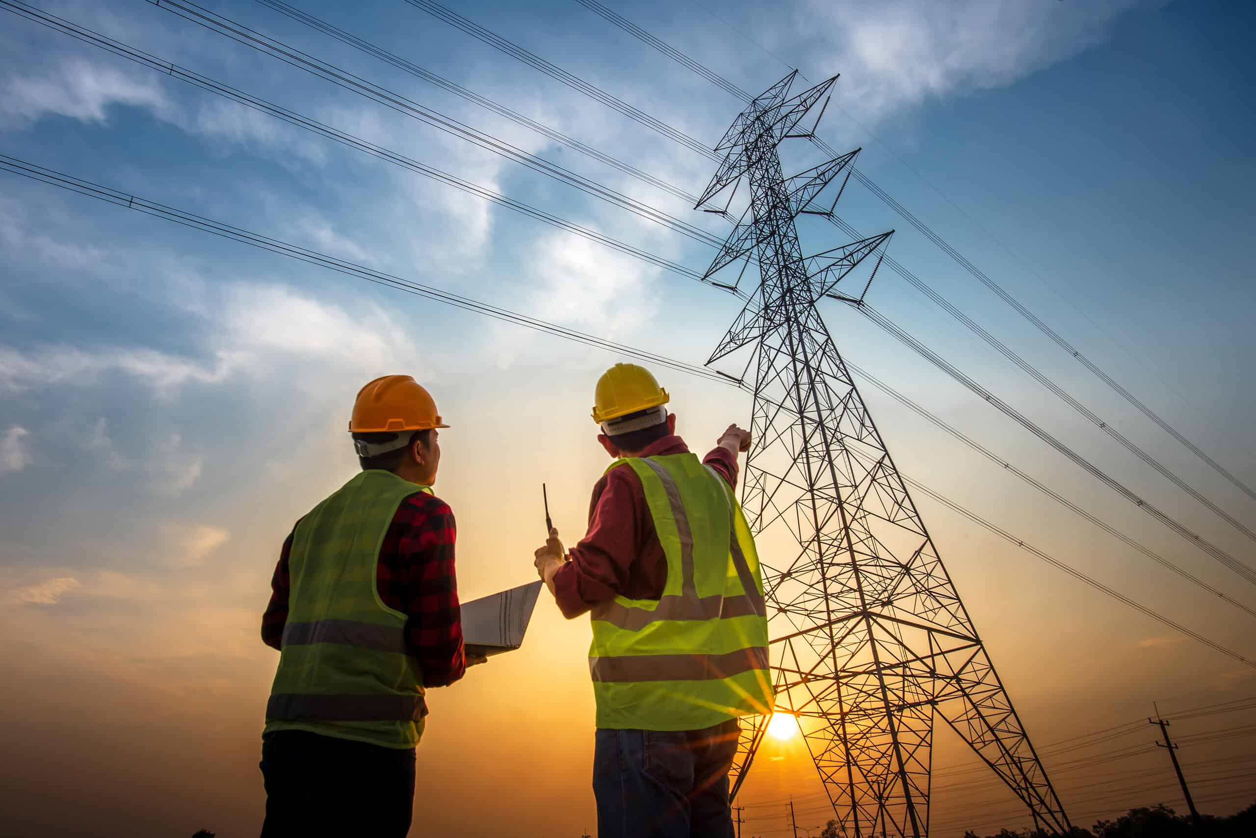 Two workers in safety vests and helmets inspect a tall power line tower at sunset, with one pointing upward and the other holding documents. The sky is partly cloudy with the sun low on the horizon. Clarisight.com