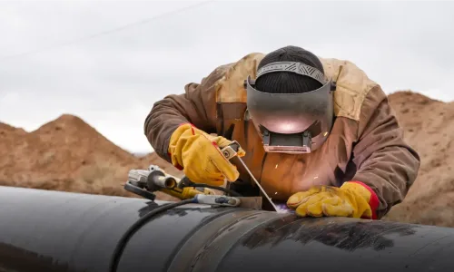 Welder in protective gear welds large pipeline outdoors.