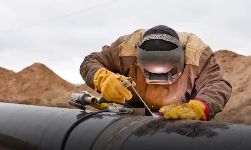 A detailed worker in safety gear welding on a pipeline outdoors, with a mountainous background, demonstrating industrial construction and maintenance.